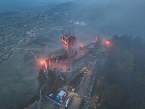 Aerial view of Guaita Tower, the historic fortress perched on Mount Titano, surrounded by mist and illuminated by warm lights in the evening in Citta di San Marino, San Marino, San Marino.