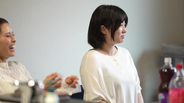 Two women share a warm kitchen moment. One laughs gently, the other looks thoughtful. Third person bends near the counter. Light spills softly over the scene. Casual, intimate, everyday moments