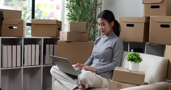Asian woman entrepreneur working on laptop, managing online orders for her small business, surrounded by cardboard shipping boxes in a home office, focusing on e-commerce growth