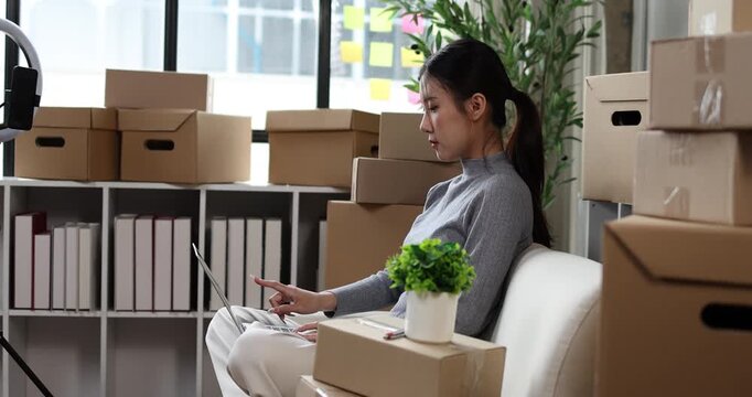 Asian woman entrepreneur working on laptop, managing online orders for her small business, surrounded by cardboard shipping boxes in a home office, focusing on e-commerce growth