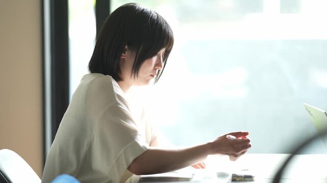 Soft light frames a focused woman at a cafe table. Her quiet concentration suggests deep thought or creative work. Natural daylight filters through the window behind her