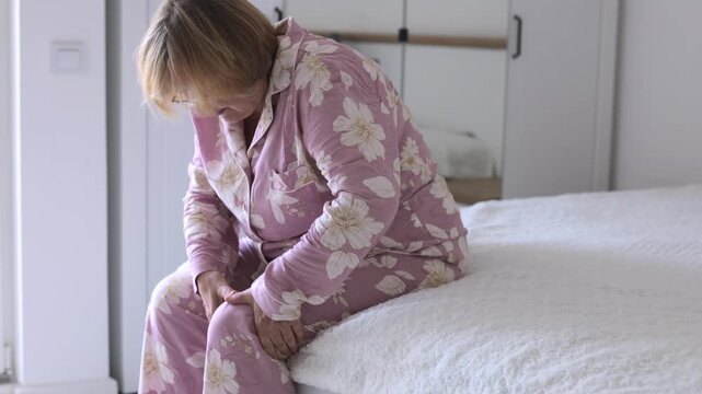Senior Woman with Knee Pain Sitting on Bed. Senior woman sitting on the edge of a bed and holding her knee at home. Mature woman in pajamas dealing with leg pain or morning discomfort.