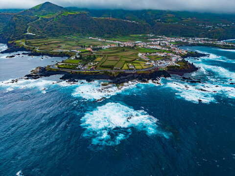 Aerial view of Mosteiros Beach with its dark volcanic sand, surrounded by lush green fields, coastal roads, and the deep blue Atlantic Ocean in Mosteiros, Acores, Portugal.