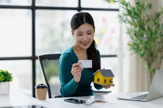 Young asian woman holding a credit card and a miniature house, planning a real estate purchase online, investing in property, and managing personal finances for home ownership