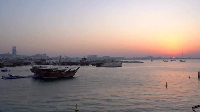 Wide sunset shot of Doha Harbor featuring traditional wooden dhow boats moored on calm reflective waters, with a glowing orange and pink sky silhouetting the city skyline in the background.
