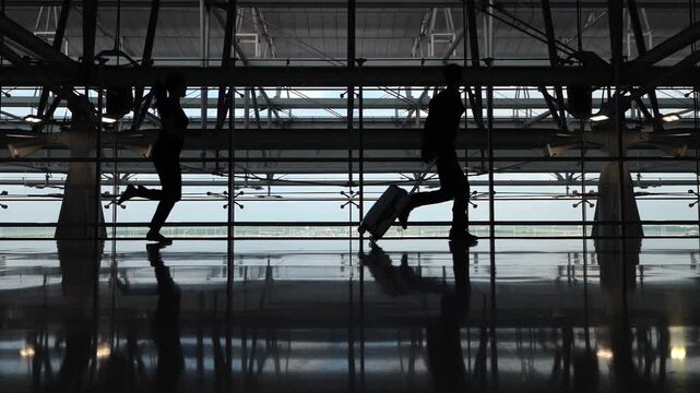 Couple of tourists run to boarding gate through empty airport terminal. They heard final call announcement and are in hurry not to miss their flight. Slow motion silhouetted shot against glass wall