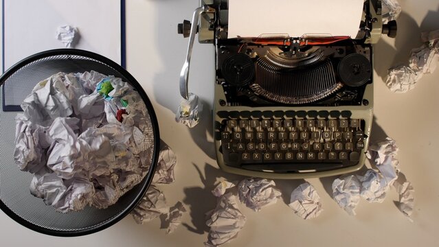 Vintage mechanical typewriter sitting on a desk with crumpled paper waste filling a nearby trash can and scattered around, symbolizing creative frustration and the challenge of idea generation