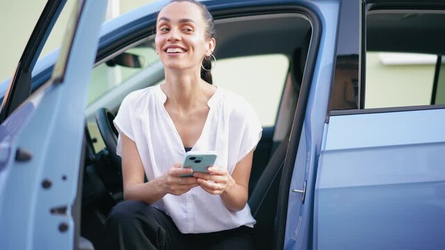 Young cheerful Hispanic woman using a smartphone while sitting in the driver&rsquo;s seat of a parked car, capturing modern lifestyle and mobile communication.