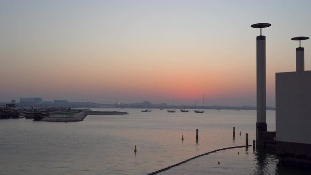 Wide sunset shot from MIA Park in Doha, Qatar, featuring calm reflective waters, silhouetted dhow boats in the harbor, structural columns in the foreground, and a warm orange glowing horizon.
