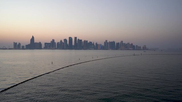 Aerial wide shot of Doha's iconic skyline at sunset, captured from the sea with calm water in the foreground and a soft orange and purple gradient sky silhouetting the modern skyscrapers.

