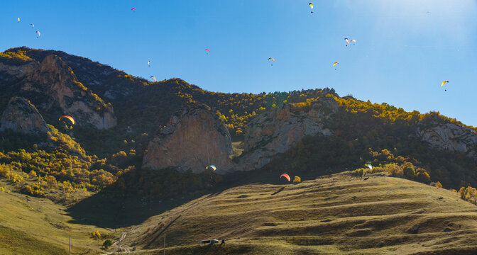 Paragliders soar gracefully against rugged mountain backdrop, with vibrant autumn foliage below and clear blue sky above. Fly Chegem paragliding site, El-Tyubu, Russia - October 7, 2024