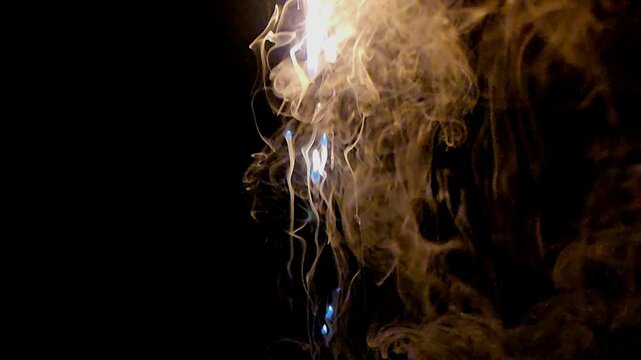 Abstract close-up footage of blue flames and swirling smoke rising from burning plastic against a deep black background. The flowing smoke creates dramatic organic patterns