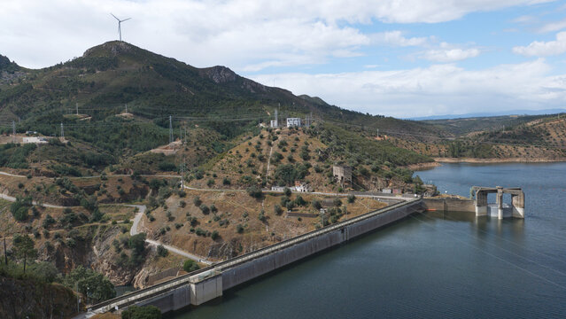 Envendos, Portugal - 23 June 2026: Aerial view of Barragem da Pracana, a concrete dam spanning the reservoir, surrounded by rolling hills, olive groves, and a wind turbine under a blue sky.
