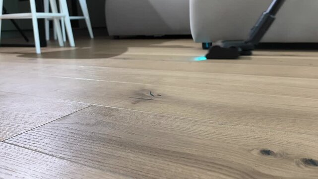 Vacuum cleaner removes dust from wooden floor in living room space. Upright device moves across parquet surface performing cleaning task. Home routine maintains hygiene and organized