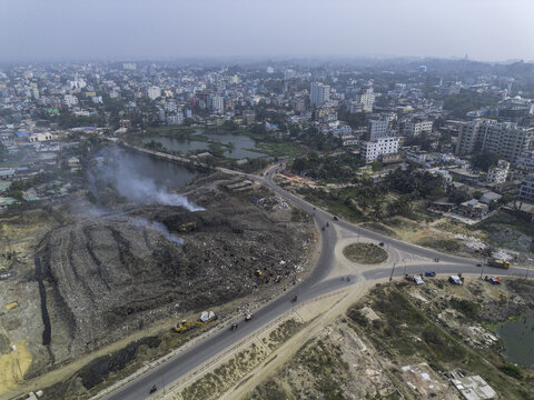 Cox's Bazar, Bangladesh - 11 March 2026: Aerial view of a large landfill site with smoke rising from waste, situated near a roundabout and urban buildings.