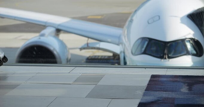 Woman pulls small silver suitcase as she walks past window in airport terminal. Shot focuses on her legs and suitcase, with blurred background showing parked plane's pilot cabin, wing, and engine.