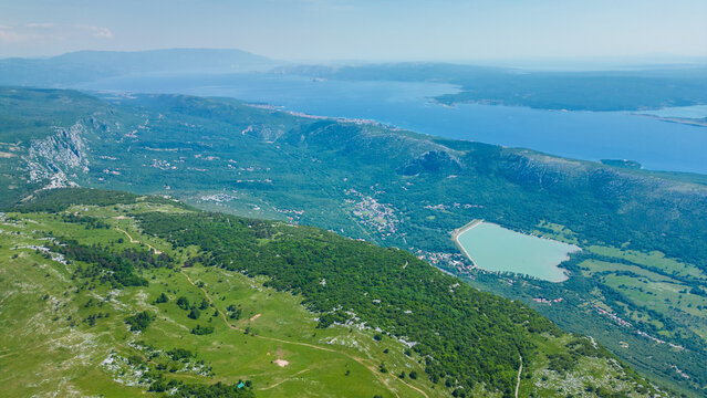 Breathtaking aerial view of Croatia's Kvarner Gulf, showcasing lush green hills, coastal towns, and the sparkling Adriatic Sea under a clear sky. A serene reservoir adds to the diverse landscape. Trib