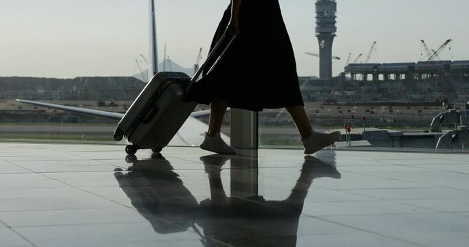 Woman walks toward boarding gate, pulling small suitcase. Her legs are reflected on smooth floor as she moves through modern terminal. In background, parked airplane and airfield