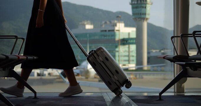 Passenger woman walks through empty airport terminal, pulling small suitcase in slow motion. Low half view captures her silhouette, focusing on her legs and luggage, as she moves past lounge window.