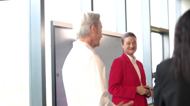 Confident young businesswoman presenting business strategy with hand drawn graph on digital whiteboard while smiling and listening to colleagues during corporate meeting in office.