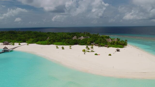 Aerial view of a Maldives resort island with crescent beach, lagoon, palm trees, thatched villas, and a long jetty as the camera glides over clear water and waves lap