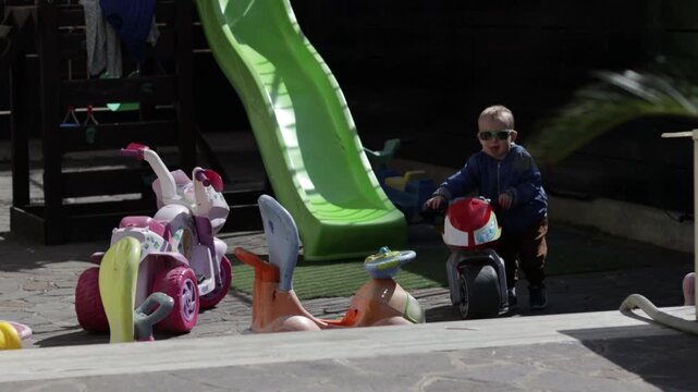 Cute toddler boy in sunglasses trying to pull a heavy plastic toy motorcycle up the steps on a playground. Little kid asking for help while playing outdoors