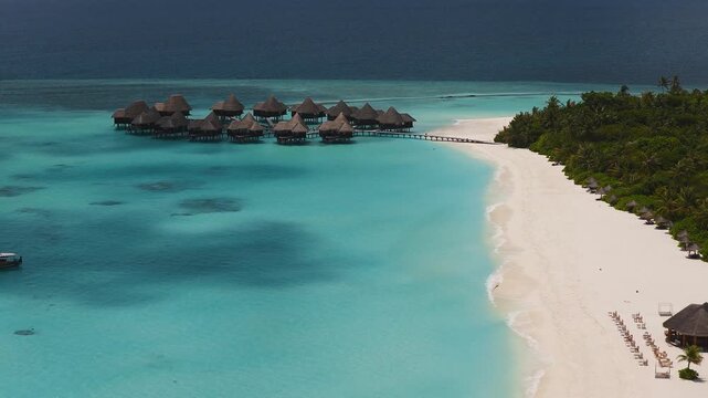 Aerial view of a Maldives resort with thatched overwater villas on stilts, a wooden jetty, palm island, sun loungers, umbrellas, and a small boat in turquoise waters.
