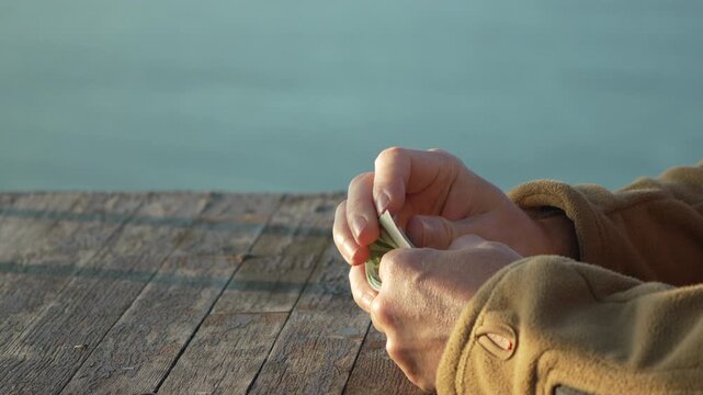 Money counting hands checking euro banknotes on wooden surface near calm water during golden hour.
