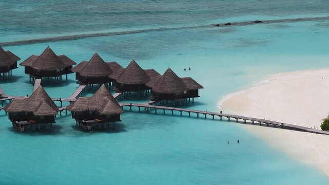 Aerial view of a Maldivian resort shows thatched villas on stilts, sinuous piers, powder white beach, vivid lagoon, reef line, swimmers, and soft waves in daylight