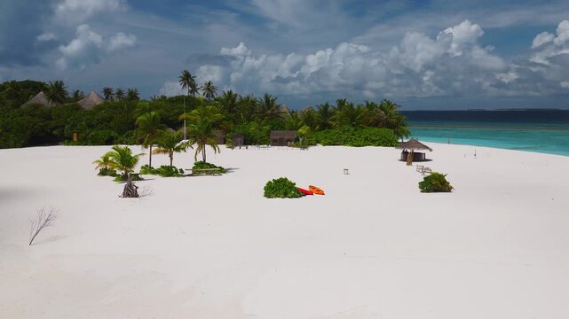 Aerial drone sweep shows Maldives island with powder white beach, coconut palms, thatch bar, low cottages, orange kayaks, reef breaks, and calm midday light