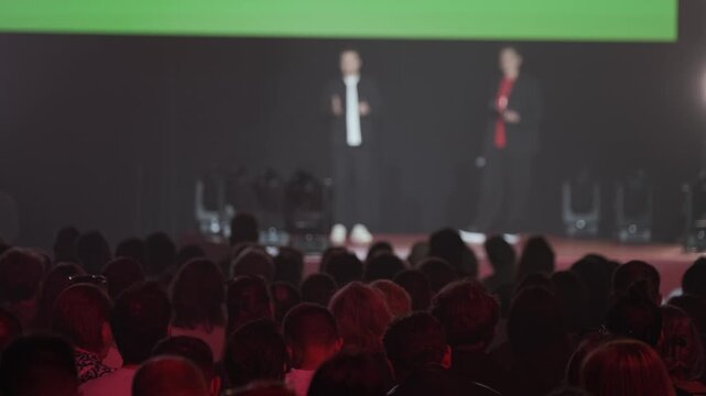 Large audience watching two speakers on stage during conference presentation, dim lighting and focused atmosphere.