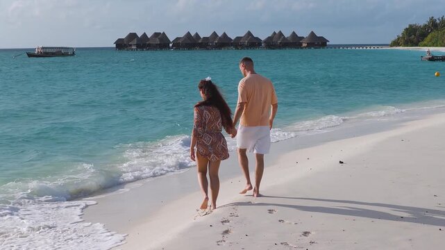 Barefoot couple walks on powder white sand as waves lap. Overwater villas on stilts and a moored dhoni drift scene at a Maldives resort in soft afternoon light.