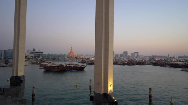 Scenic dusk shot framed between MIA Park columns, overlooking Doha Corniche harbor with traditional dhow boats, the glowing Fanar spiral minaret, and city skyline under a soft pink sky.
