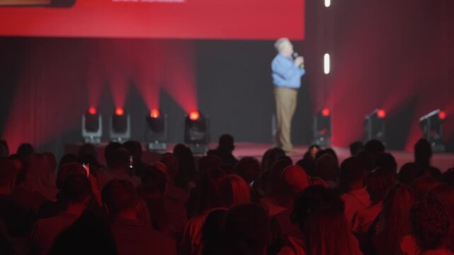 Conference speaker on stage addressing large audience, red stage lighting and projection screen, people watching and recording on phones, corporate event atmosphere.