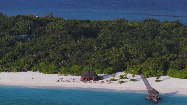 Aerial Maldives resort on a small atoll shows pale sand, a beachfront pavilion, a wooden jetty, a boat at the pier, palms, a mast, gentle surf, guests, and warm light.