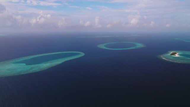 Aerial view of ring shaped coral atolls and a sandy islet in the Maldives, with surf along rims and the deep Indian Ocean, as the camera makes a sweeping pass.