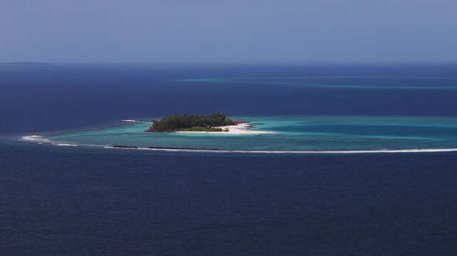 Aerial view of a Maldivian atoll island with white sand, palm canopy, shallow turquoise lagoon, deep ocean, clear reef break, and a faint pier, camera slowly tracks