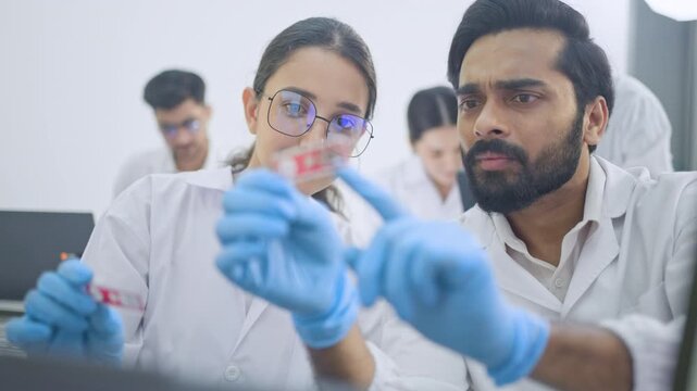 Young woman in eyeglasses holding specimen while bearded man looks doubtfully, research team discussion in busy laboratory, focus on sample.