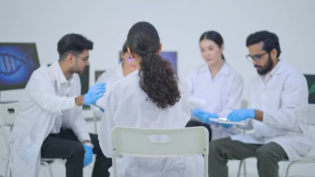 Group of five scientists in lab coats sitting together comparing notes on tablets and printouts, DNA image on screen, science team briefing.