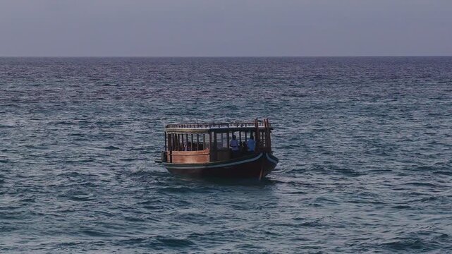 Aerial dhoni with two crew moves toward camera on turquoise sea near a palm island and atoll rim in the Maldives, shot wide in soft late afternoon light