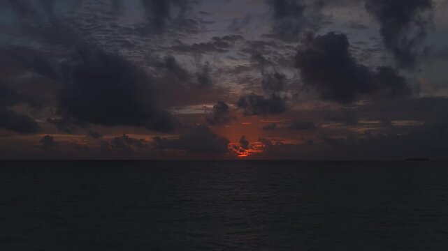 Aerial wide shot at dusk shows Maldives open water, a tiny atoll at far right, heavy cumulus and rippled clouds, orange sunset band, drifting clouds, gentle sea ripples.