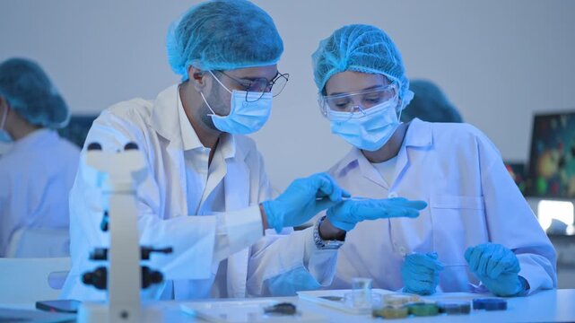 Two scientists in protective caps and masks gesturing during discussion at lab bench with microscope, night research in blue light.