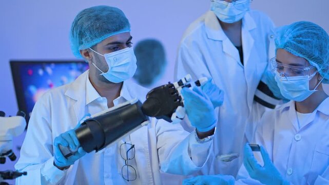 Male engineer in mask and hairnet examining black robotic prosthetic hand with colleagues, bionics and medical technology development.
