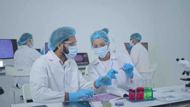 Male and female researchers in masks examining samples at bench with test tube rack and digital tablets, busy research laboratory team.