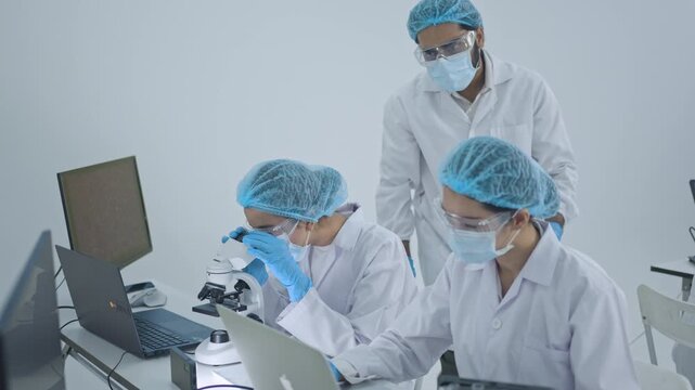 Scientist looking through microscope while colleague supervises and woman works on laptop, research team analyzing samples in modern lab.