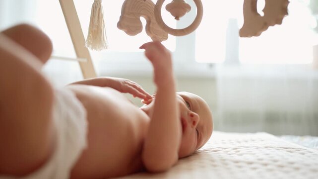 Curious newborn baby boy in a diaper lying on a bed and playing with a wooden mobile toy arch, exploring the world and developing his senses and fine motor skills in a cozy nursery