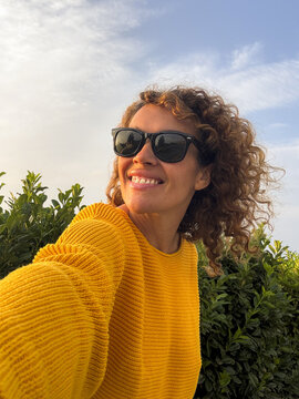 Smiling woman taking a selfie outdoors wearing sunglasses and a yellow sweater with curly hair against green foliage and a blue sky