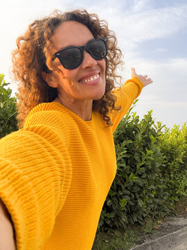 A smiling woman in a yellow sweater takes an outdoor selfie with sunglasses, arms outstretched, enjoying sunshine and nature near green bushes