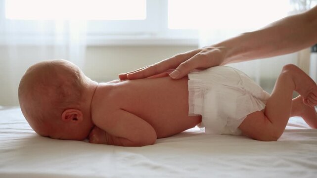 Caring mother's hand gently massaging a newborn baby's back. An infant in a diaper is doing tummy time on a bed, strengthening muscles and developing coordination through physical therapy