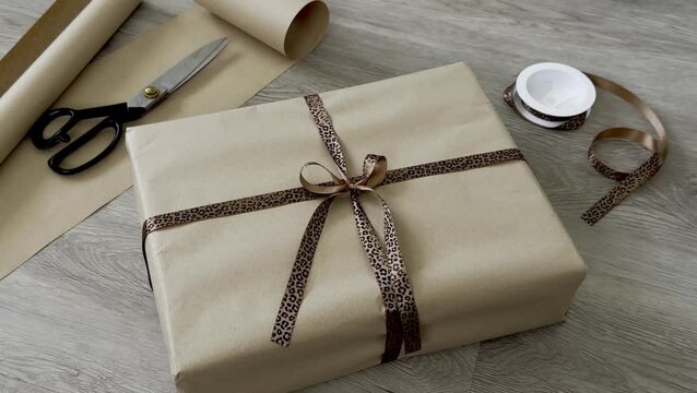 Gift-wrapped package with brown paper and leopard print ribbon, surrounded by scissors, wrapping paper roll, and tape on a wooden surface